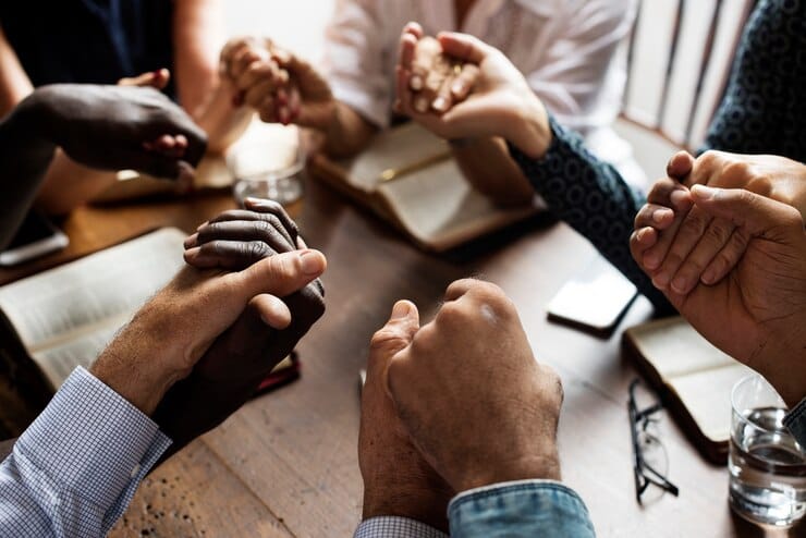 Grupo de pessoas segurando as mãos em um momento de oração e conexão espiritual, com Bíblias abertas sobre a mesa.