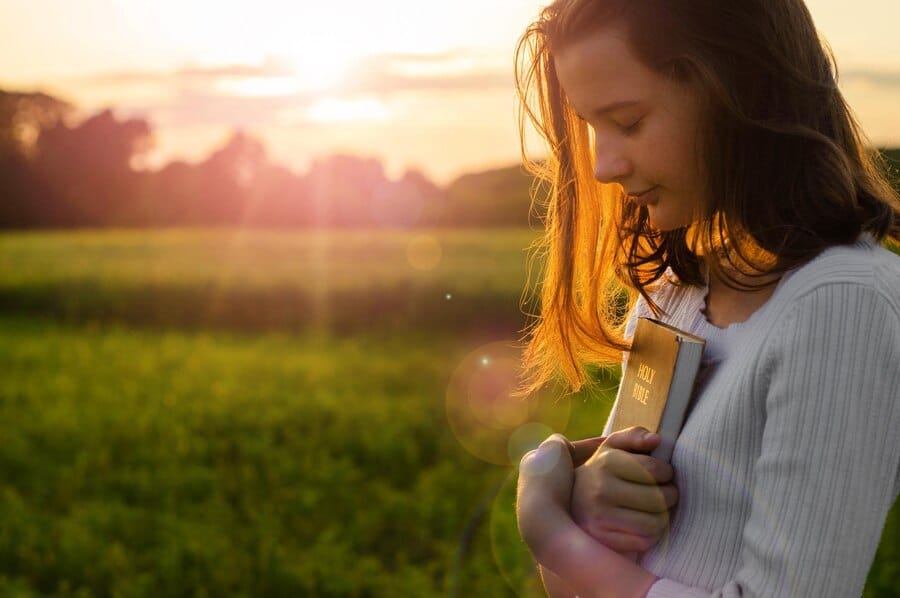 "Imagem de uma adolescente em pé, com a cabeça baixa e segurando uma Bíblia, em um momento de oração ou gratidão. A cena representa entrega, fé e confiança nos planos de Deus para sua vida.
