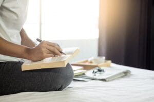 Imagem de uma mulher em sua cama, com uma caneta na mão e escrevendo em um livro, simbolizando um momento de preparação espiritual. A cena transmite introspecção, conexão com a fé e busca por sabedoria divina.