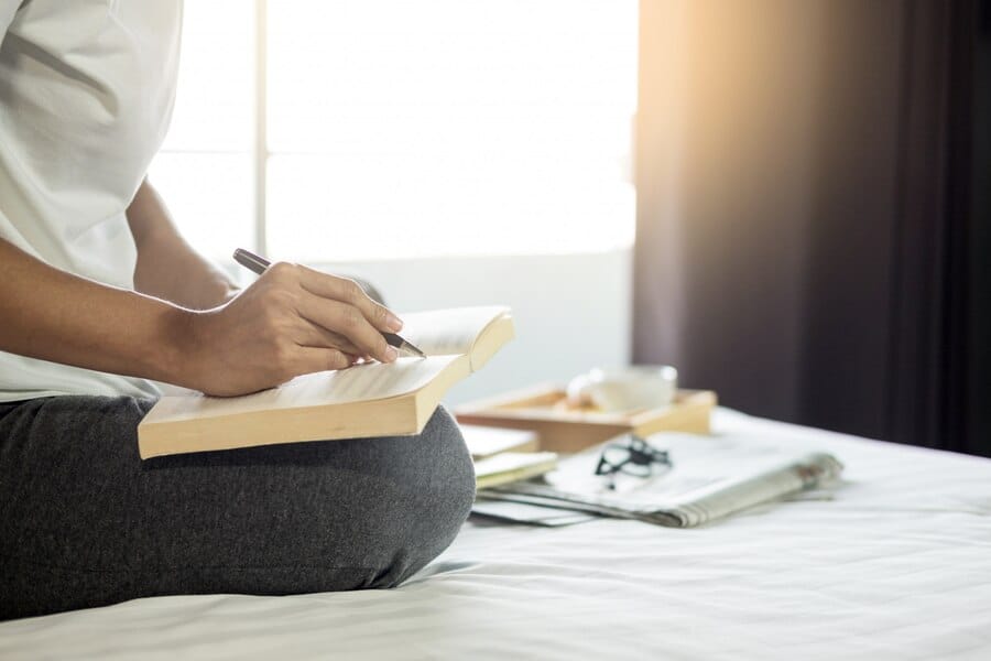 Imagem de uma mulher em sua cama, com uma caneta na mão e escrevendo em um livro, simbolizando um momento de preparação espiritual. A cena transmite introspecção, conexão com a fé e busca por sabedoria divina.
