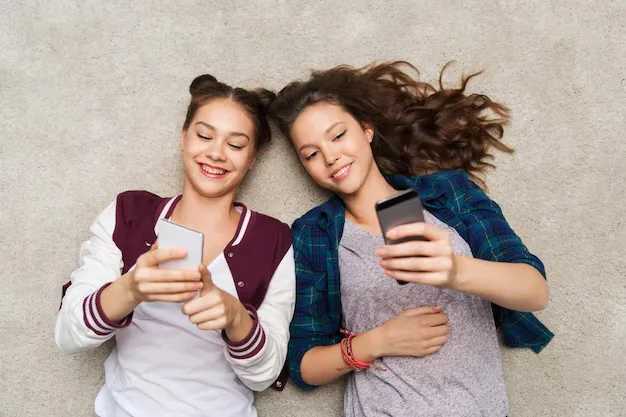Dois jovens deitadas no chão, sorrindo e interagindo com seus celulares, transmitindo diversão e conexão digital.