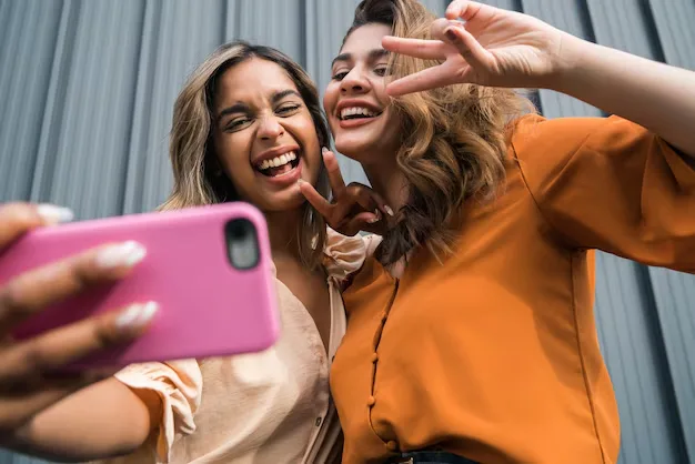 Duas amigas tirando uma selfie animada com um celular rosa, fazendo poses divertidas e expressando alegria.