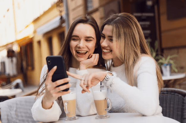 Duas amigas em um café rindo e olhando para um celular enquanto tomam café com leite, usam suéteres brancos.