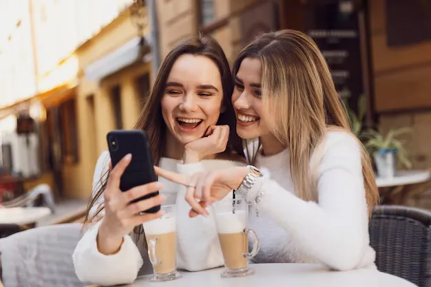 Duas amigas em um café rindo e olhando para um celular enquanto tomam café com leite, usam suéteres brancos.