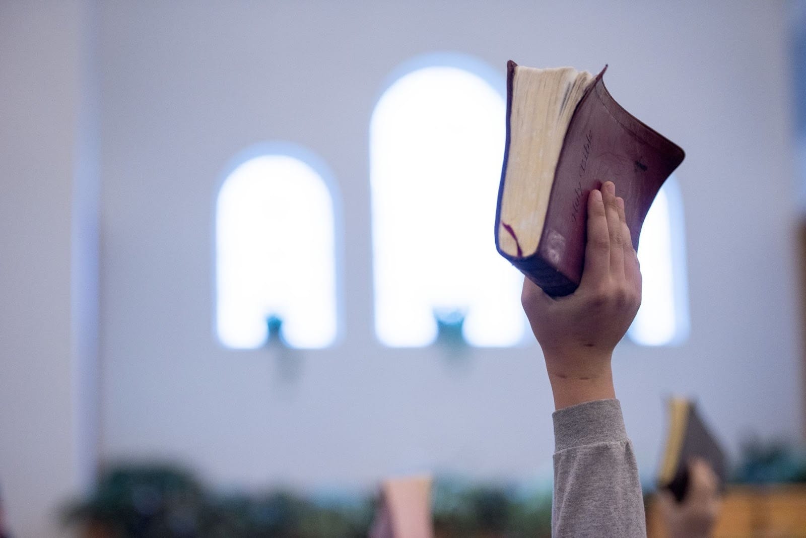 Em um close-up, um braço esticado, vestindo uma manga cinza, segura uma Bíblia de capa bordô no alto. O livro está virado com as páginas para cima. O fundo é uma parede de igreja branca, com duas ou mais janelas em arco desfocadas e iluminadas. A imagem tradicional de adoração, com a Bíblia levantada, contrasta com a palavra-chave, sugerindo o potencial da Inteligência Artificial nas igrejas para ampliar ou apoiar atos de fé e pregação.