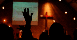 Silhuetas de pessoas em um ambiente escuro e quente, durante um culto de adoração. Em primeiro plano, uma mão está levantada. Ao fundo, uma tela de projeção exibe letras (possivelmente de louvor), e uma cruz iluminada em vermelho se destaca em uma parede de tijolos. A imagem ilustra a adoração e o ambiente de gravação de Conteúdo Cristão.