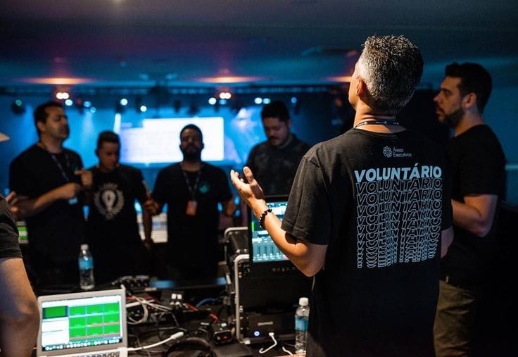 Visão de costas de um voluntário em uma equipe de multimídia ou bastidores (com uma camisa que diz "Voluntário") em um evento. Ele está de pé perto de um console de áudio ou vídeo, de braços abertos, em oração, junto a outros membros da equipe (homens) em um ambiente escuro de show ou conferência, antes de produzir Conteúdo Cristão ou iniciar um culto/evento.