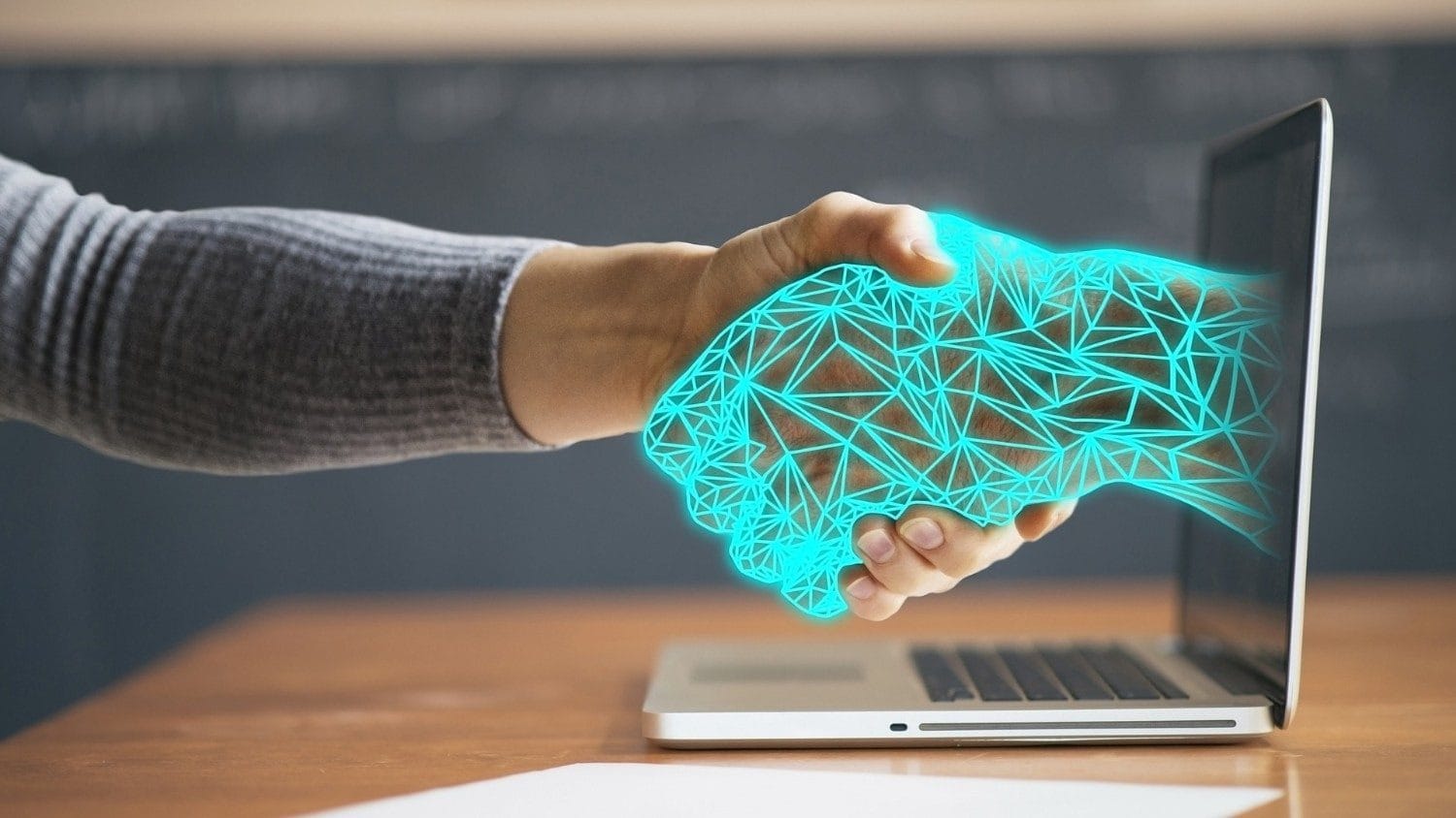 Uma mão humana, vestindo uma manga cinza, aperta a mão de uma mão digital futurista em neon azul claro, que se estende para fora da tela de um notebook prateado aberto. A cena se passa sobre uma mesa de madeira em um ambiente de sala de aula ou escritório. A imagem simboliza a colaboração, a confiança ou o aperto de mão entre a humanidade (representada pela igreja/fé) e a Inteligência Artificial nas igrejas (representada pela tecnologia).